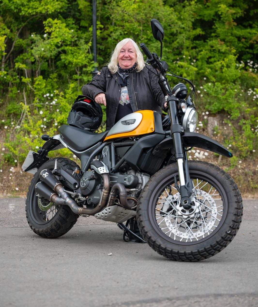 Maxine and her bike Woman with shoulder-length platinum-blonde hair leaning against her yellow and black Scrambler motorbike, wearing a biker jacket and jeans.